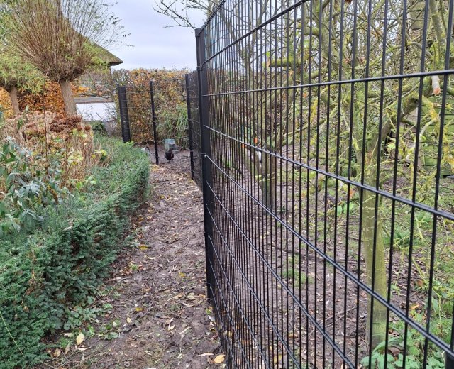 Zwart enkelstaafmat hekwerk in tuinomgeving met zichtbare begroeiing en onderhoudspad